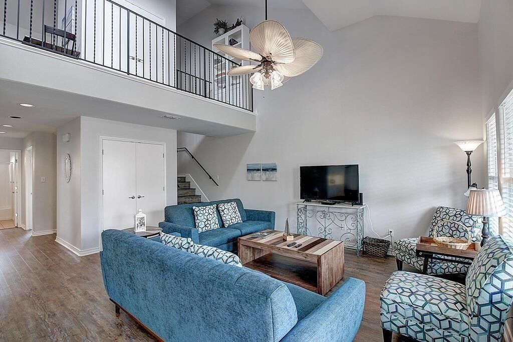A spacious living room with a high ceiling, a large ceiling fan, a flat-screen TV, and a staircase leading to an upper level.