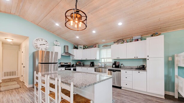 A modern kitchen with white cabinetry, a central island with bar stools, stainless steel appliances, and a decorative clock. - A- Rocks Resort by Silver Sands Vacation Rentals (Port Aransas)