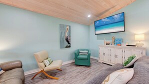A living room with a wooden ceiling, a teal armchair, a beige sofa, a grey bed, and a white cabinet with decorative items. - A- Rocks Resort by Silver Sands Vacation Rentals (Port Aransas)