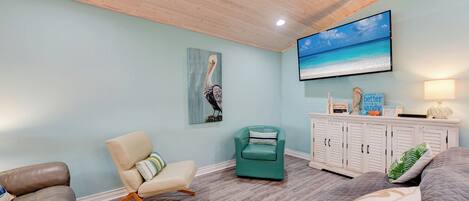 Un salon avec un plafond en bois, un fauteuil turquoise, un canapé beige, un lit gris et un meuble blanc agrémenté d’objets décoratifs.