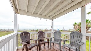 A covered porch with wooden flooring and white railings, featuring three Adirondack chairs and string lights. - A- Rocks Resort by Silver Sands Vacation Rentals (Port Aransas)