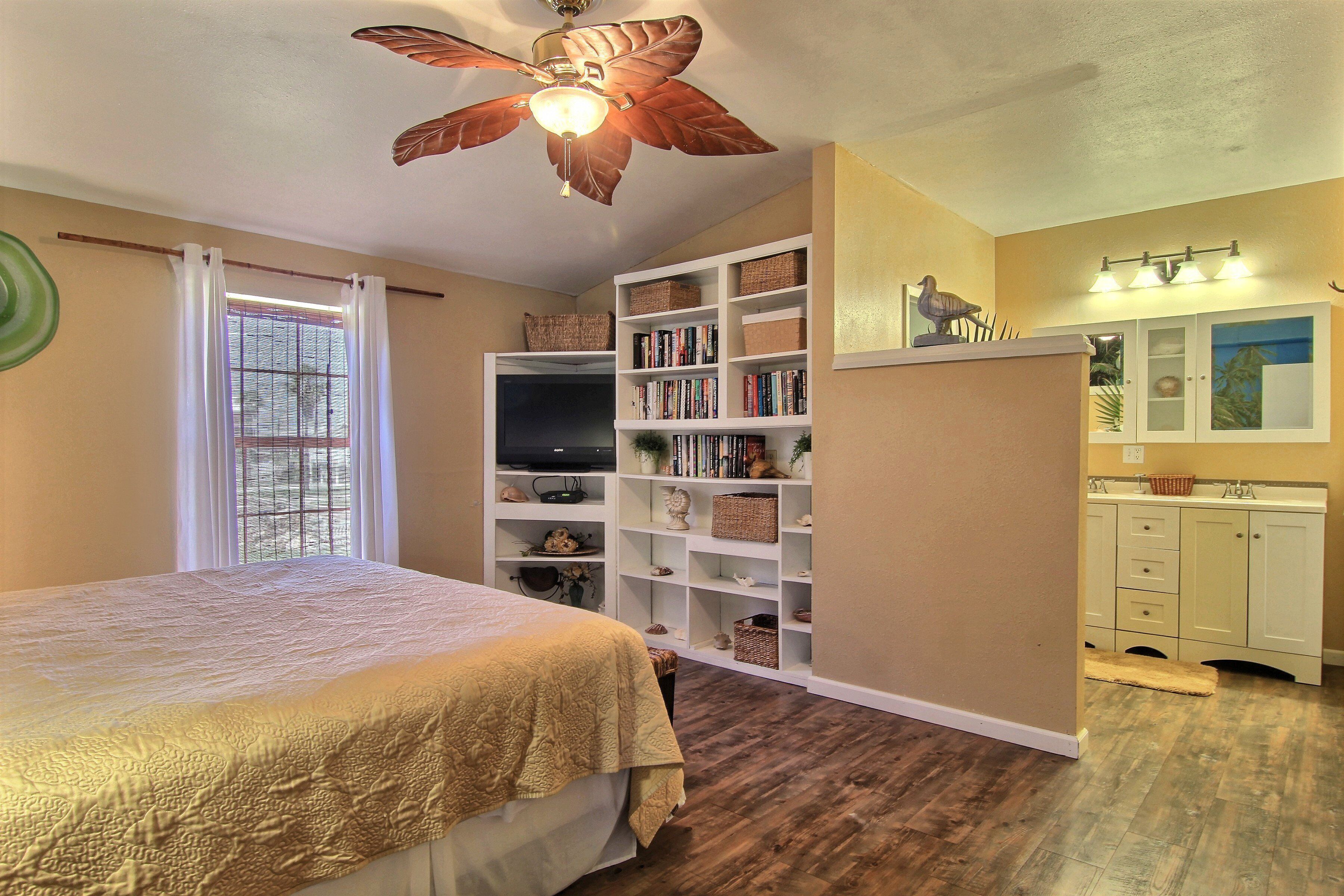 A bedroom with a bed, a ceiling fan, a bookshelf, and a bathroom with a sink and mirror.