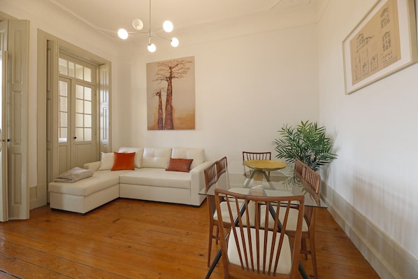 A living room with a white sofa, a glass coffee table, wooden chairs, a large window, and a painting on the wall.