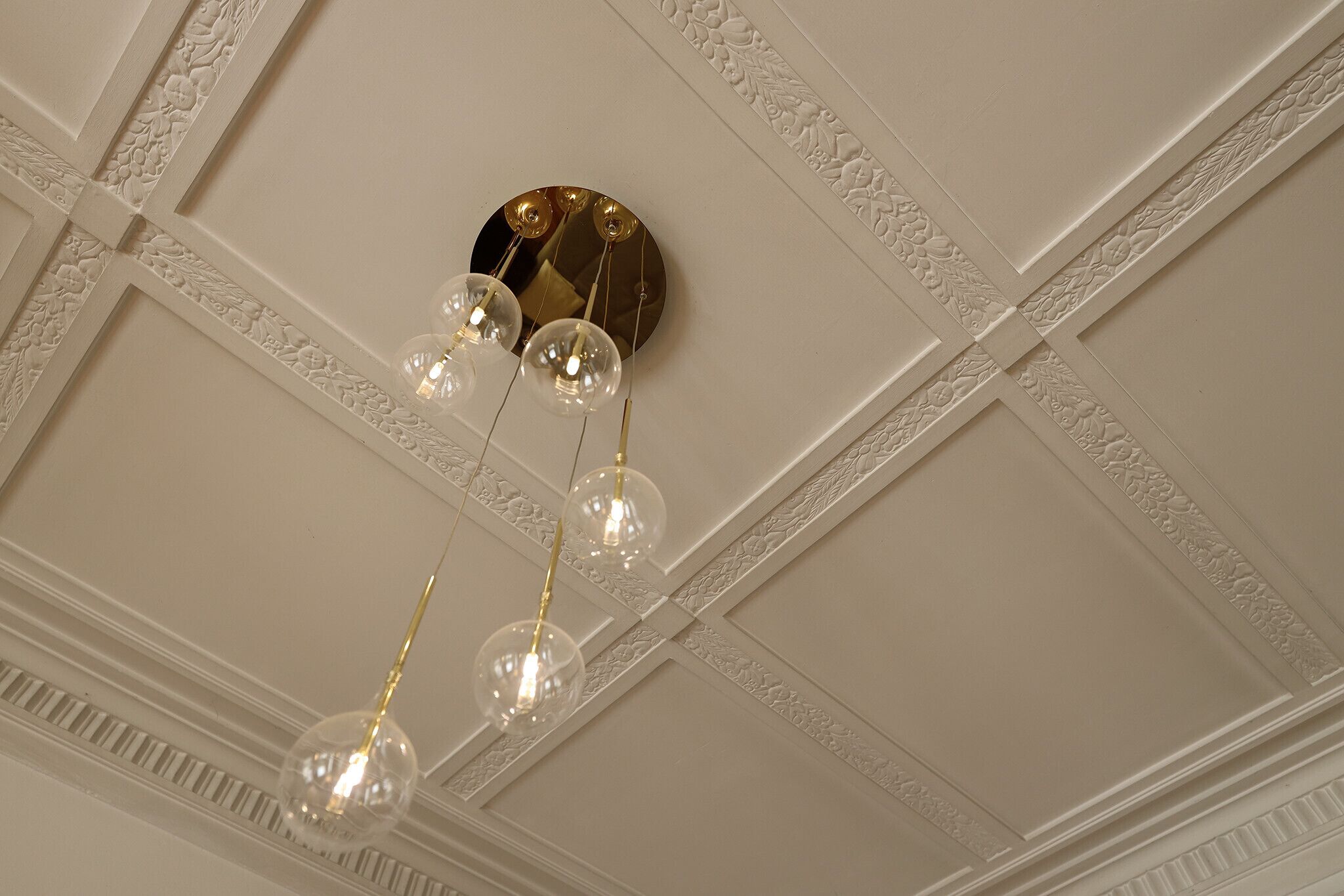 Un plafond orné de moulures décoratives et un lustre composé de globes en verre transparent et d’éléments dorés.