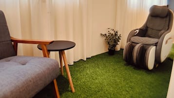 A room with a grey sofa, a small round table, a massage chair, and a potted plant.