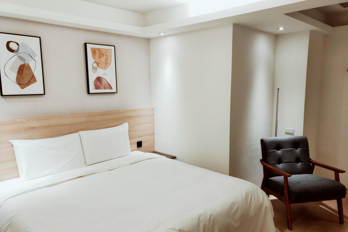A hotel room with a large bed, a wooden headboard, two framed artworks on the wall, and a dark upholstered chair.