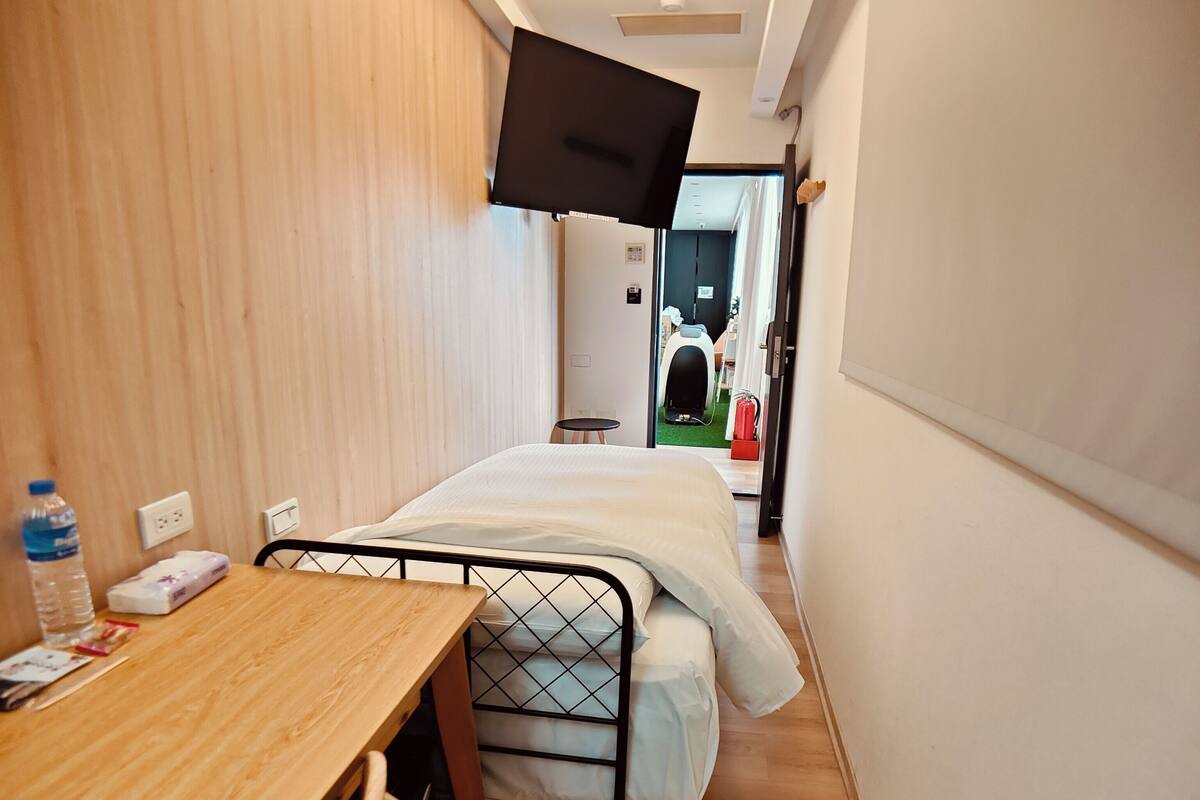 A narrow hotel room with a wooden desk, a bed with a metal frame, and a flat-screen TV mounted on the wall.