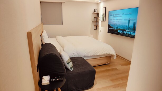 A compact bedroom with a bed, a small seating area, a TV mounted on the wall, and a shelf with decorative items. - FooYee Hotel-YongHe (New Taipei City)