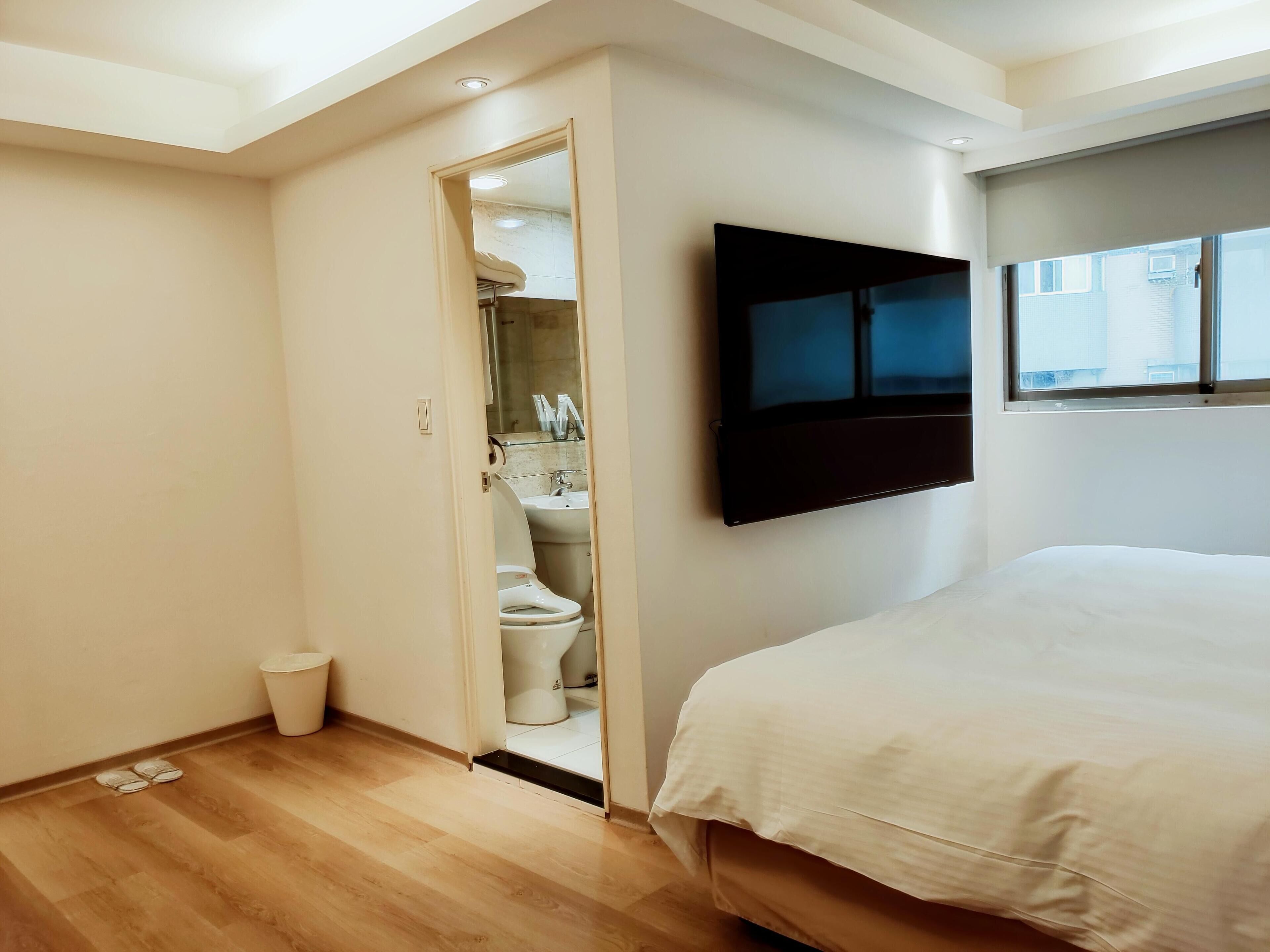 Une chambre à coucher moderne, dotée d’un grand lit, d’un téléviseur fixé au mur et d’une salle de bain visible à travers une porte ouverte.