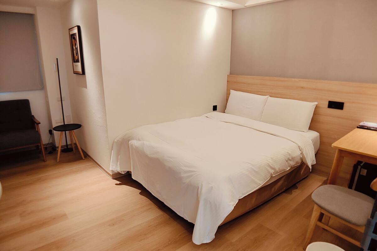 A modern hotel room with a large bed, a small desk, a chair, and a trash bin.