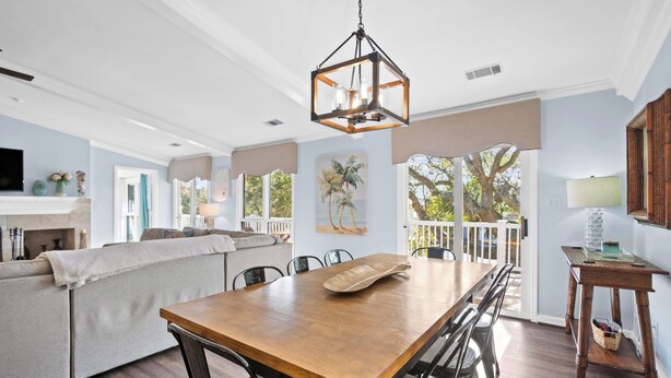 A spacious living area with a wooden dining table, black chairs, a beige sofa, and a large window offering a view of trees and a balcony. - FrippLife, Walk to Beach, Golf Cart Included - 707 Bluefish (Fripp Island)