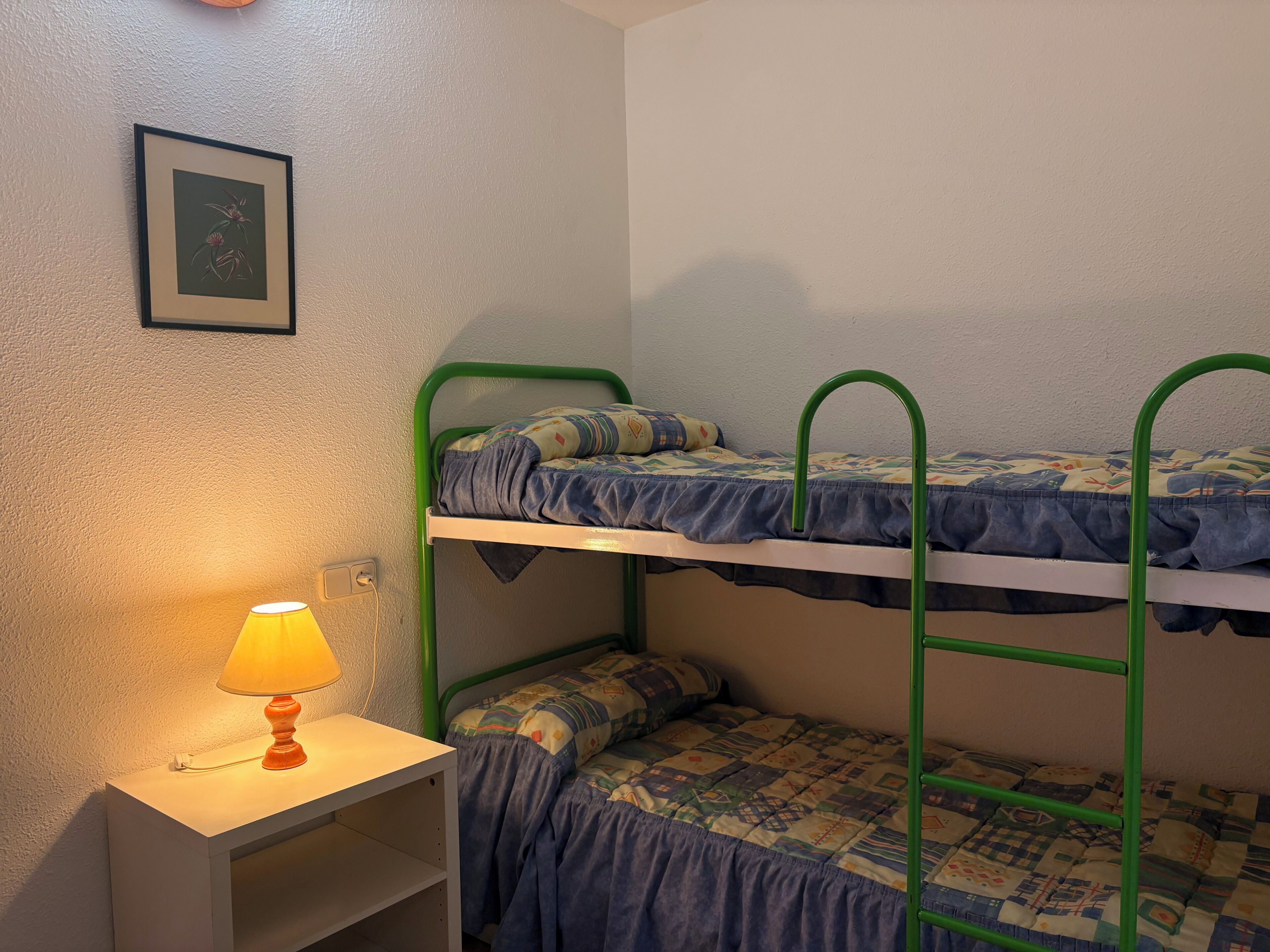 Ein Etagenbett mit grünem Gestell, ein Nachttisch mit Lampe und ein gerahmtes Bild an der Wand.