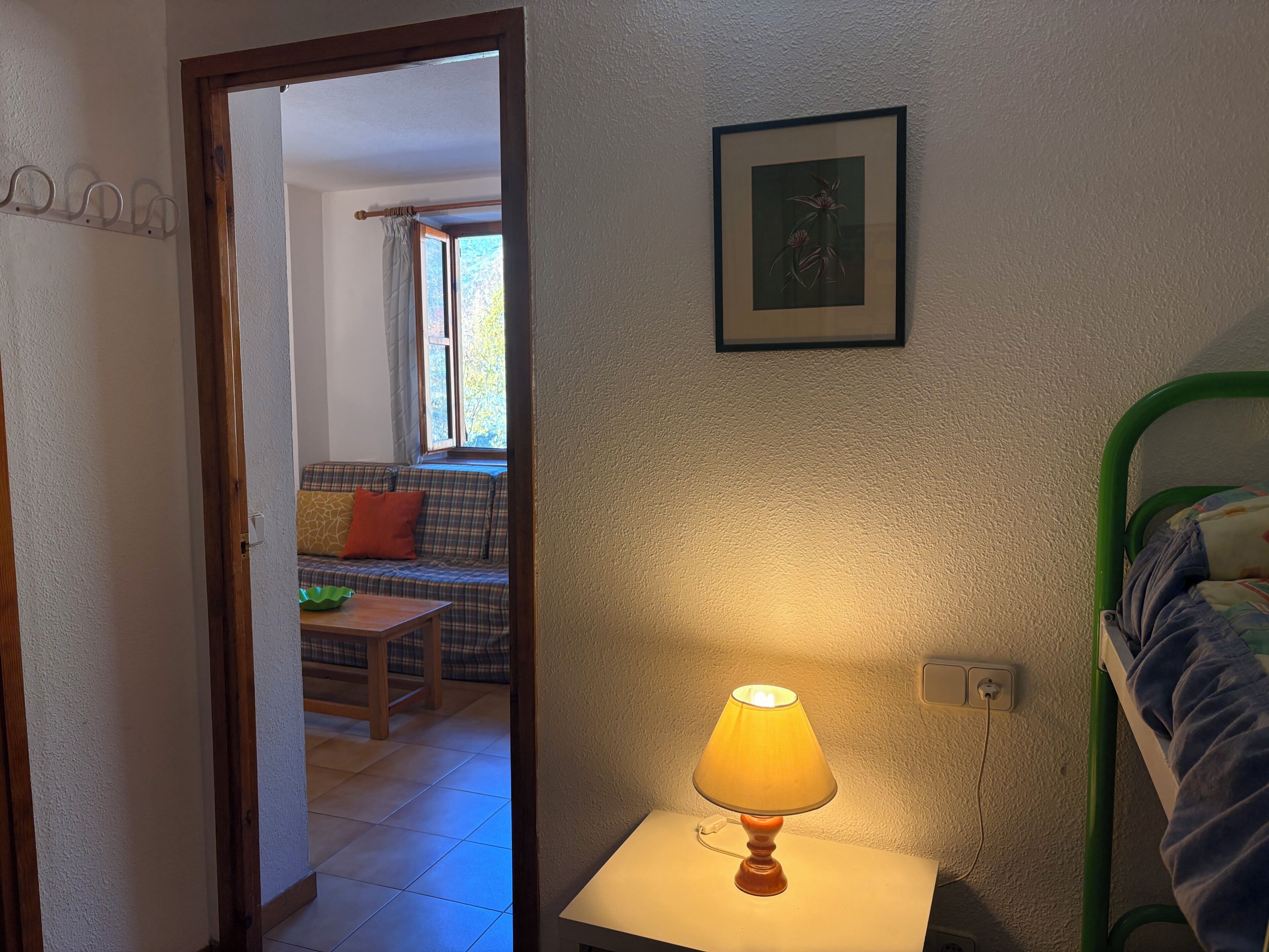 Ein Zimmer mit einem Bett, einer Lampe auf einem Nachttisch, einem gerahmten Bild an der Wand und Blick auf einen Wohnbereich mit einer Couch und einem Fenster.