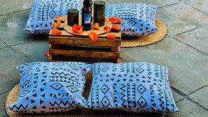 Una mesa de madera con botellas y pétalos de flores, rodeada de cojines estampados sobre un suelo de concreto.