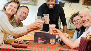 Un grupo de personas brindando con vasos, sentadas en una mesa con mantel a cuadros. Se ve una caja con la etiqueta 'KUMILTY' sobre la mesa.