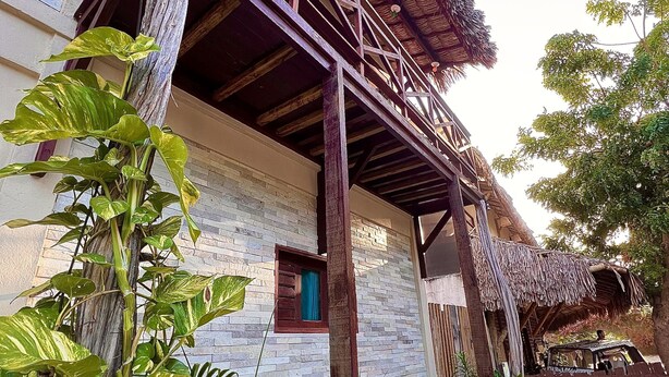 A traditional wooden structure with a thatched roof, a small window with blue curtains, and lush green plants in front. - Residencial Bem te vi (Jijoca de Jericoacoara)
