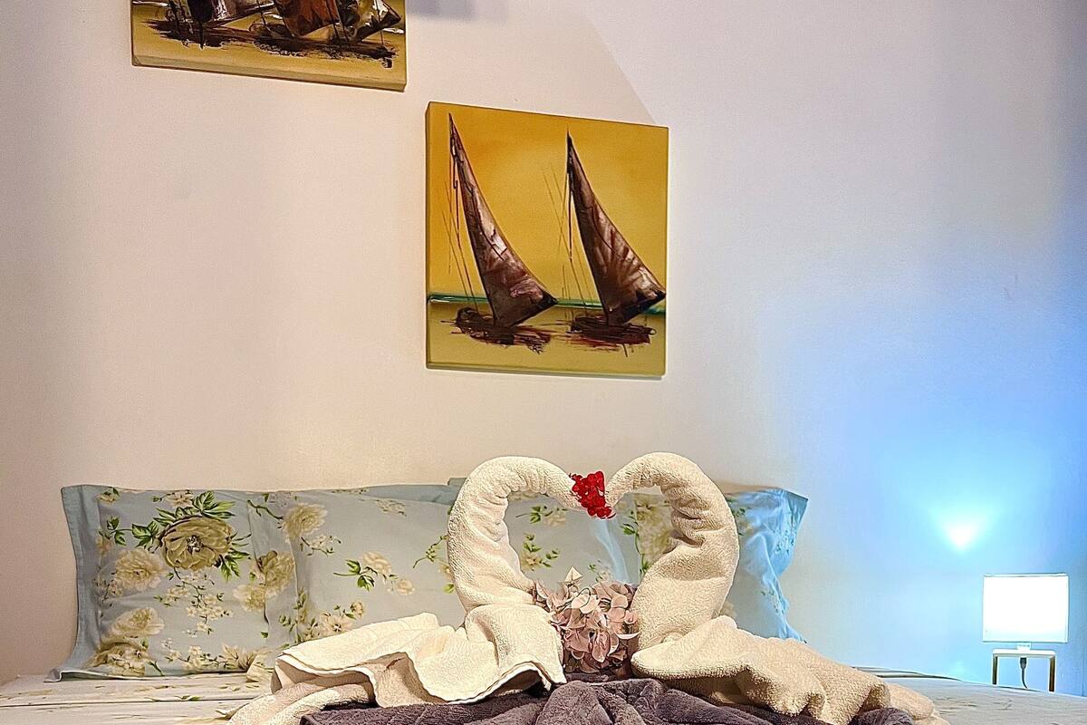 A neatly made bed with a floral bedspread, a heart-shaped towel arrangement, and two sailboat paintings on the wall.