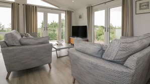 A spacious living room with two grey patterned sofas, a flat-screen TV, and large windows offering a view of greenery. - 7 Faraway Fields (Liskeard)