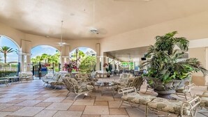 A covered outdoor seating area with chairs and tables, surrounded by potted plants and a view of a building in the background.