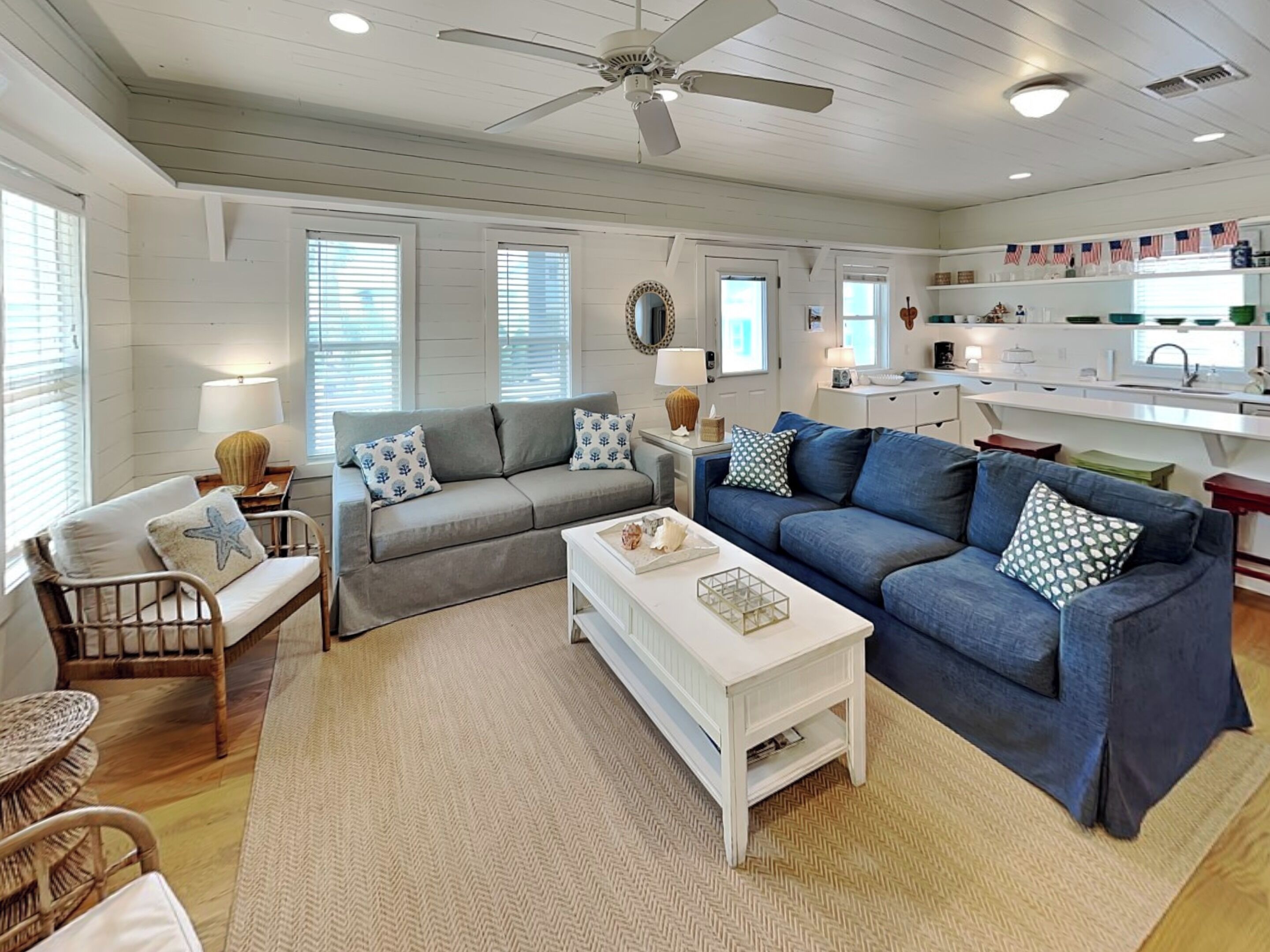 A spacious living room with a grey sofa, blue sofa, and a white coffee table. There is a ceiling fan, a kitchen area with white cabinets, and a dining area with wicker furniture.