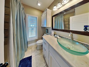 A bathroom with a glass sink, a toilet, a shower curtain, and a window with blinds. - Baja Breeze by Silver Sands Vacation Rentals (Port Aransas)