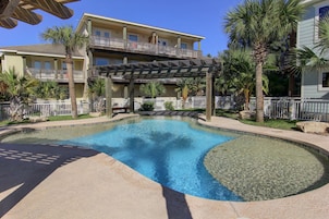 A swimming pool with a wooden pergola, palm trees, and a multi-story building with balconies.