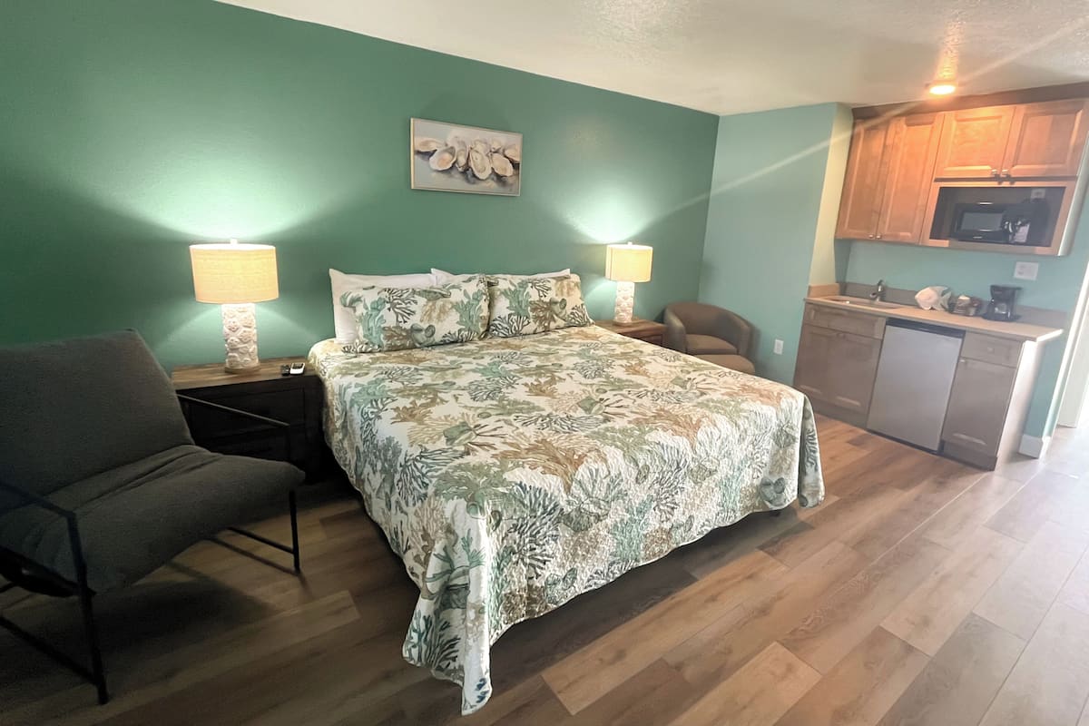 A bedroom with a bed, bedside lamps, a chair, a small desk, and a kitchenette.