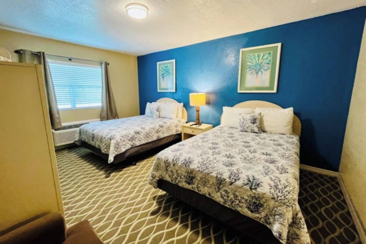 A hotel room with two beds, a window with blinds, a nightstand with a lamp, and two framed pictures on the wall.