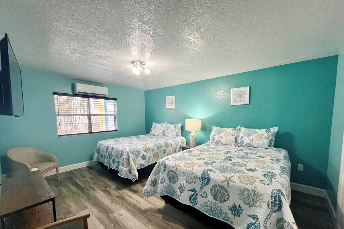 A room with two beds, a desk, a chair, a window with blinds, and a ceiling fan.
