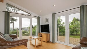 Amplia sala de estar con sofá de cuero, mesa de centro de madera y televisor de pantalla plana. Grandes puertas corredizas de vidrio dan a una terraza exterior con vistas a zonas verdes.
