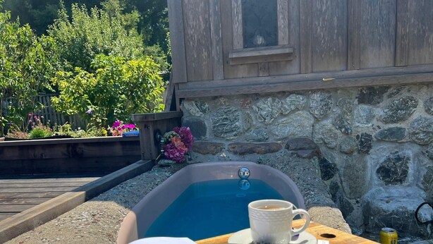 A wooden deck with a stone wall, a hot tub, a cup of coffee, and an open book. - Mendocino Coast Home with Sauna and Fireplace  (Fort Bragg)