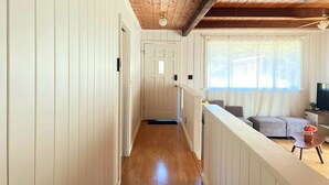 A hallway with wooden floors and white walls, leading to a living area with a sofa, a TV, and a window with curtains. - Mendocino Coast Home with Sauna and Fireplace  (Fort Bragg)