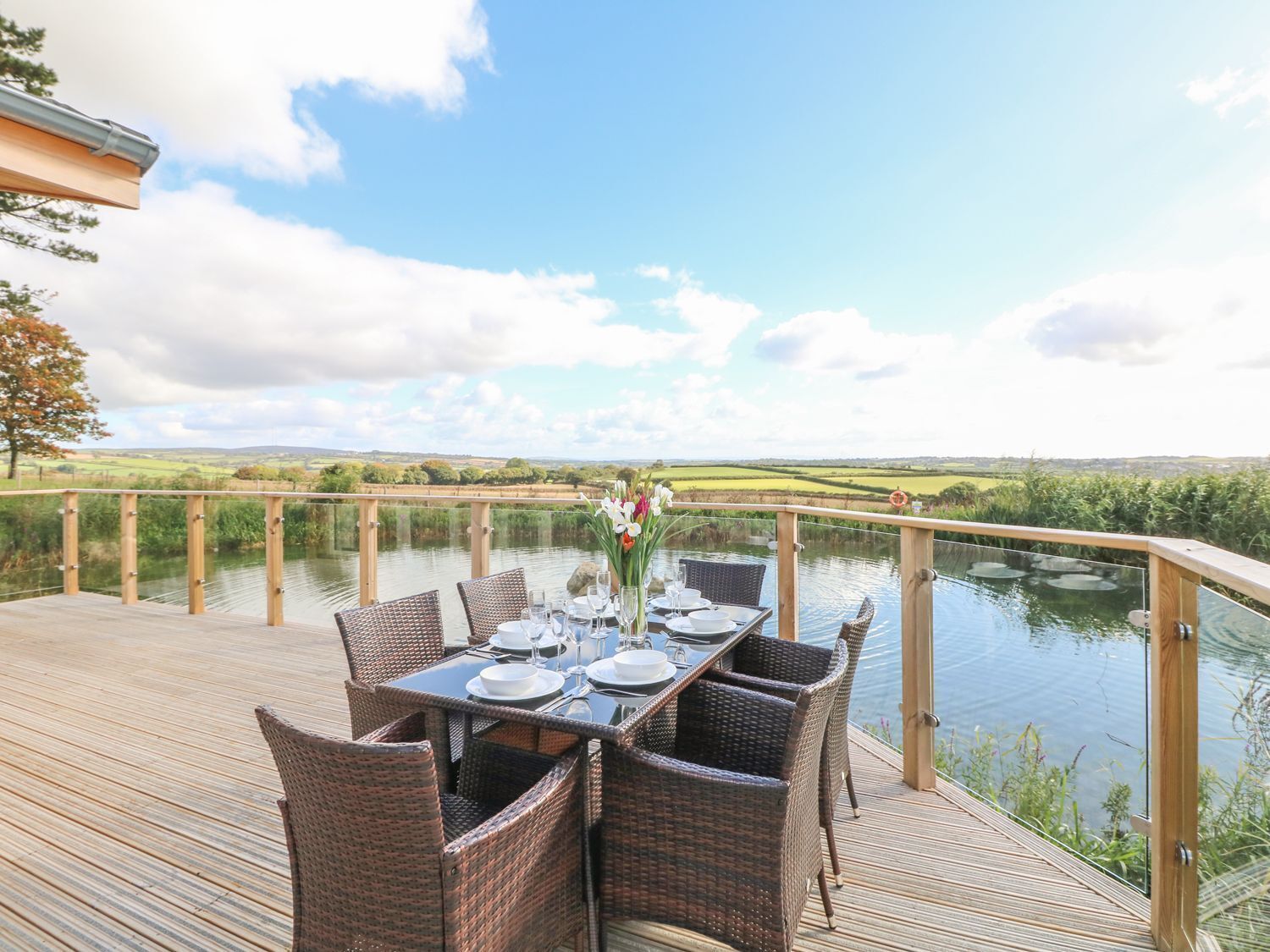 Mesa de comedor lista para dos en una terraza de madera con sillas de mimbre, con vista a un cuerpo de agua tranquilo y un paisaje pintoresco.