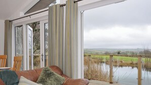 Ein gemütliches Zimmer mit einem braunen Ledersofa, gestreiften Vorhängen und Blick auf einen See und Felder.