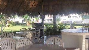 An outdoor patio area with a round table and white plastic chairs, a grill, and a white fence in the background. - CONDO MARINA G  BY TRVL2HM (Nuevo Nayarit)