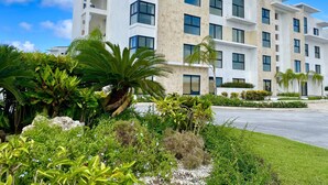 Un hôtel moderne à plusieurs étages, doté d’une façade blanche, de grandes fenêtres et d’un jardin bien entretenu où poussent différentes espèces de plantes, ainsi qu’un palmier.
