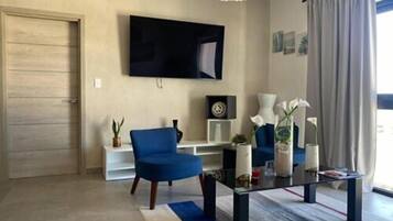 A modern living room with a flat-screen TV mounted on the wall, a blue armchair, a glass coffee table, and a large window with curtains.