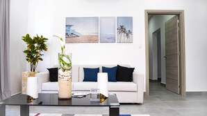 Un salon moderne avec un canapé, une table basse en verre et un vase rempli de fleurs. Trois tableaux encadrés ornent le mur, et une porte donne accès à une autre pièce.
