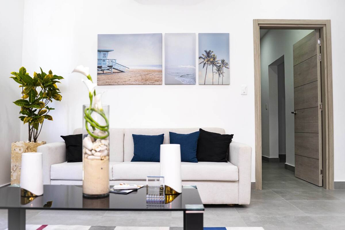 A modern living room with a sofa, a glass coffee table, and a vase with flowers. There are three framed pictures on the wall, and a door leading to another room.