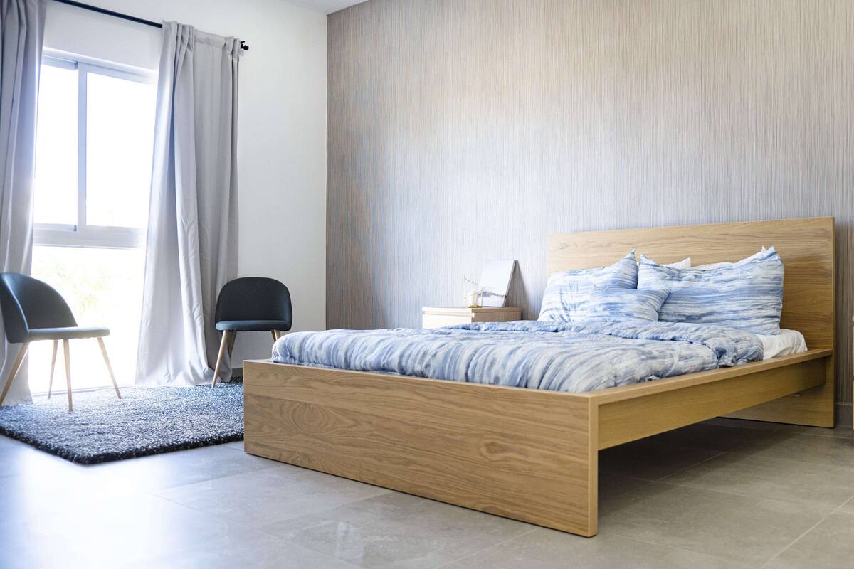A modern bedroom with a wooden bed, a nightstand, a chair, and a view of the outdoors through a window with curtains.