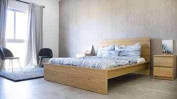 A modern bedroom with a wooden bed, a nightstand, a chair, and a view of the outdoors through a window with curtains.