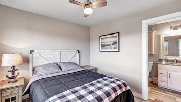 Een slaapkamer met een bed, een nachtkastje, een plafondventilator, een ingelijst schilderij aan de muur, en een badkamer zichtbaar door een geopende deur.