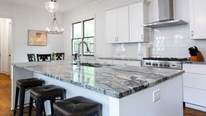 A modern kitchen with white cabinets, a large island with bar stools, a stainless steel range hood, and a pendant light.