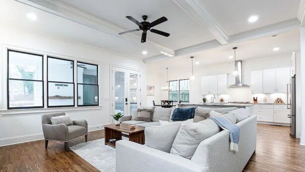 A spacious living room with a large sectional sofa, a coffee table, and a grey armchair. There is a kitchen in the background with white cabinets and stainless steel appliances.