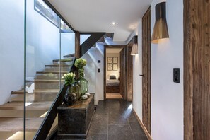Uma escadaria moderna com guarda-corrimão de vidro, um banco de madeira com objetos decorativos e uma luminária que emana luz quente.