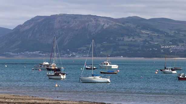 A coastal scene with multiple sailboats anchored near a beach, with a mountainous backdrop. - No 62 (Beaumaris)