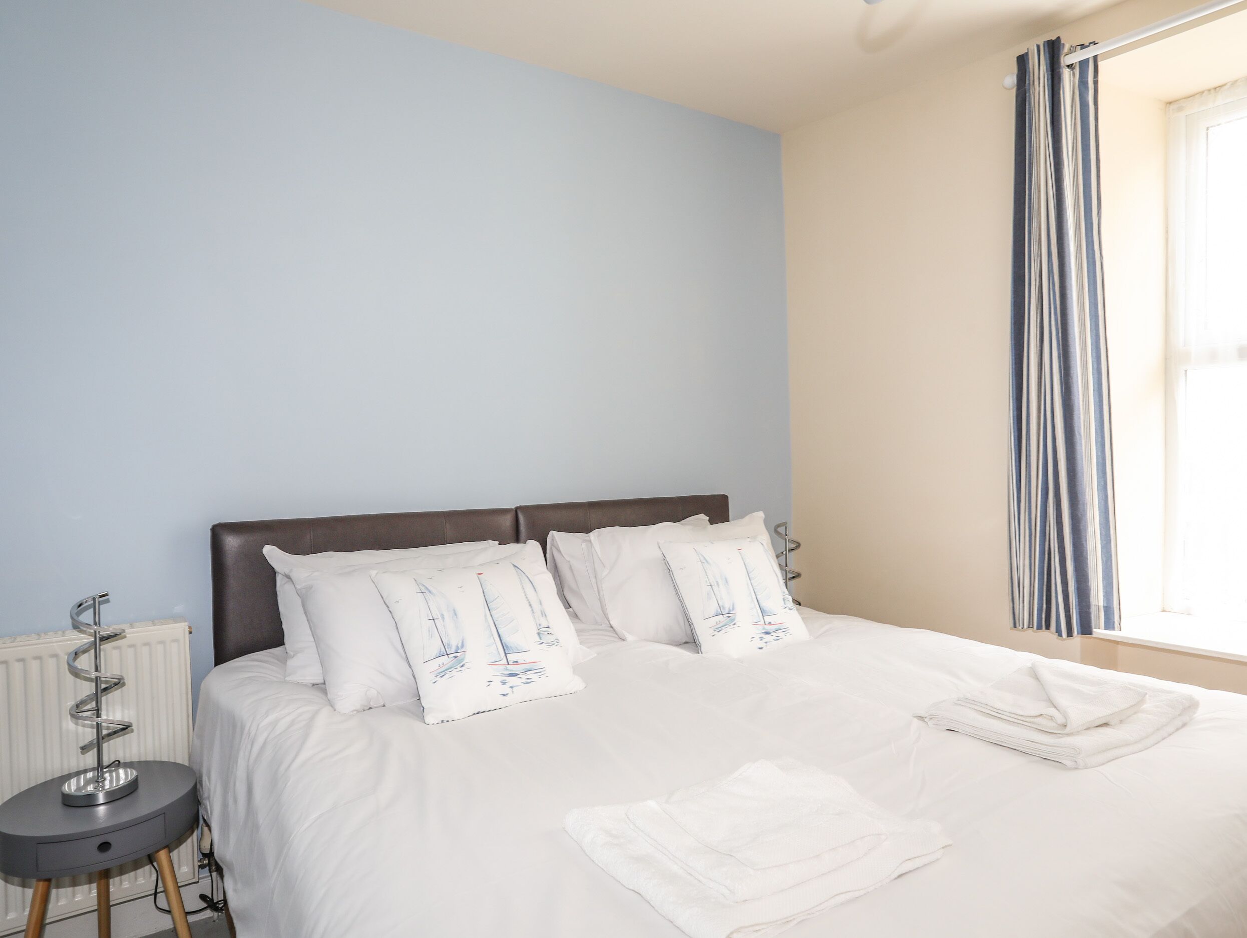 A neatly made bed with white bedding and pillows, a small bedside table with a lamp, and a window with curtains.
