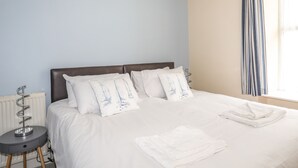 A neatly made bed with white bedding and pillows, a small bedside table with a lamp, and a window with curtains. - No 62 (Beaumaris)