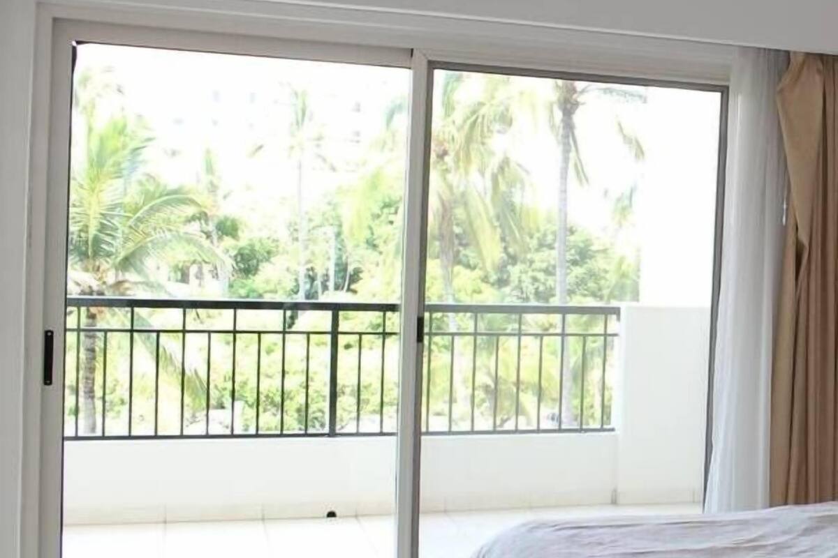 A bedroom with a bed, a ceiling fan, and a sliding glass door leading to a balcony with a view of palm trees.