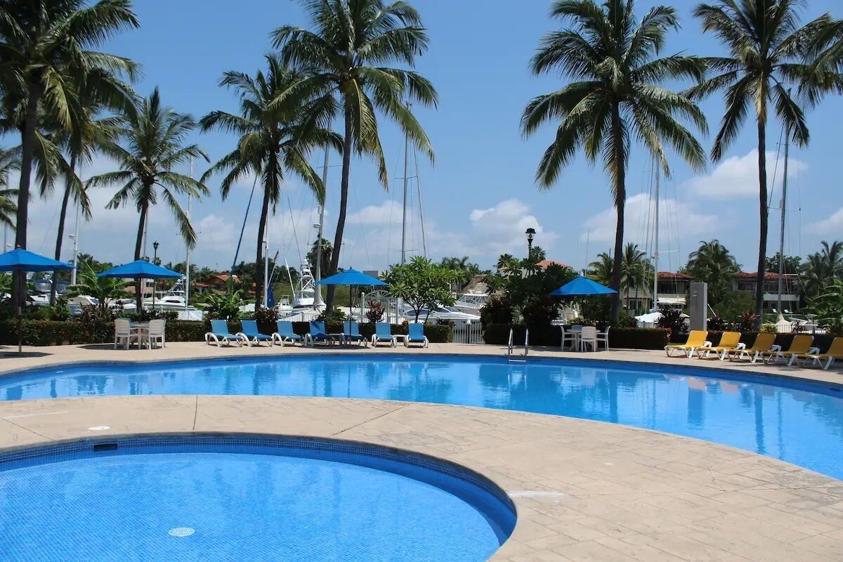 A pool with lounge chairs and umbrellas, surrounded by palm trees.